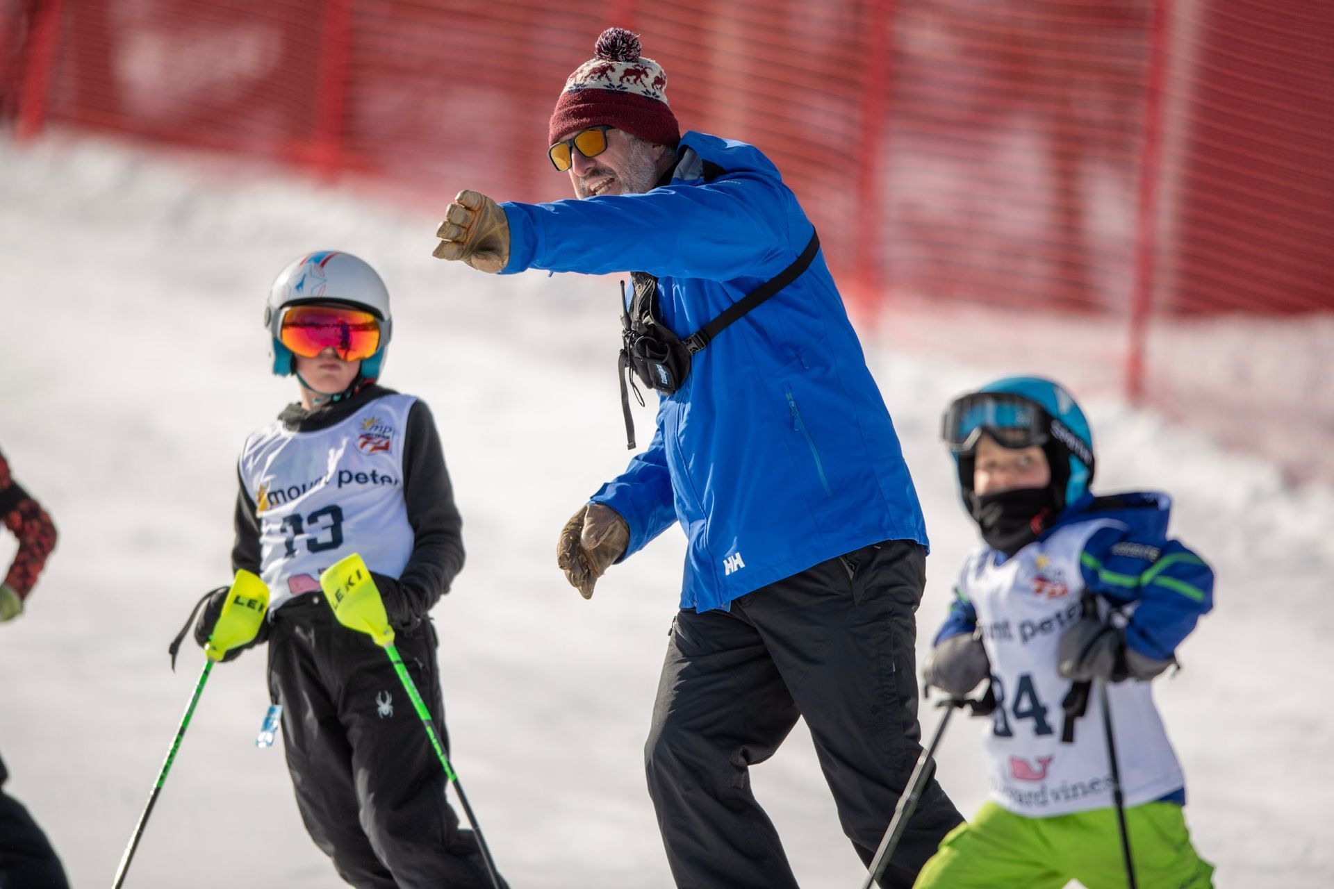 Coach gesturing to young skiers on a snowy slope. The children wear ski gear and goggles.