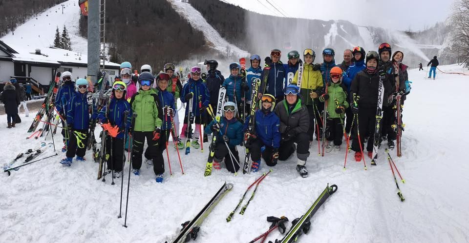 Group of skiers on a snowy slope, posing with skis and poles. Mountains and buildings in the background.
