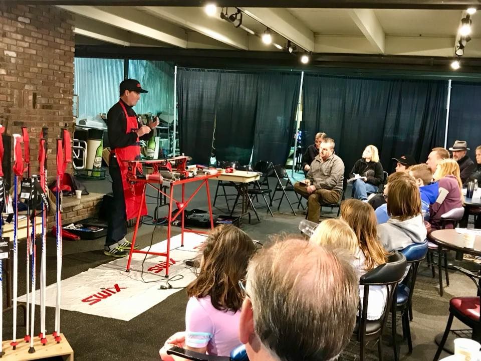 Man in red apron demonstrates ski repair to a seated audience in a room with skis and equipment.