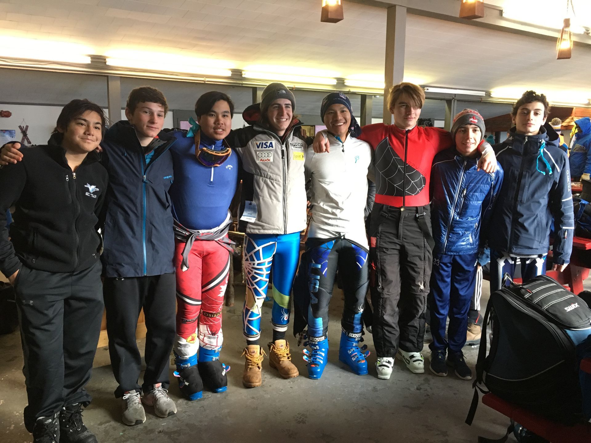 A group of skiers in colorful racing gear and ski boots, arm in arm, standing indoors.