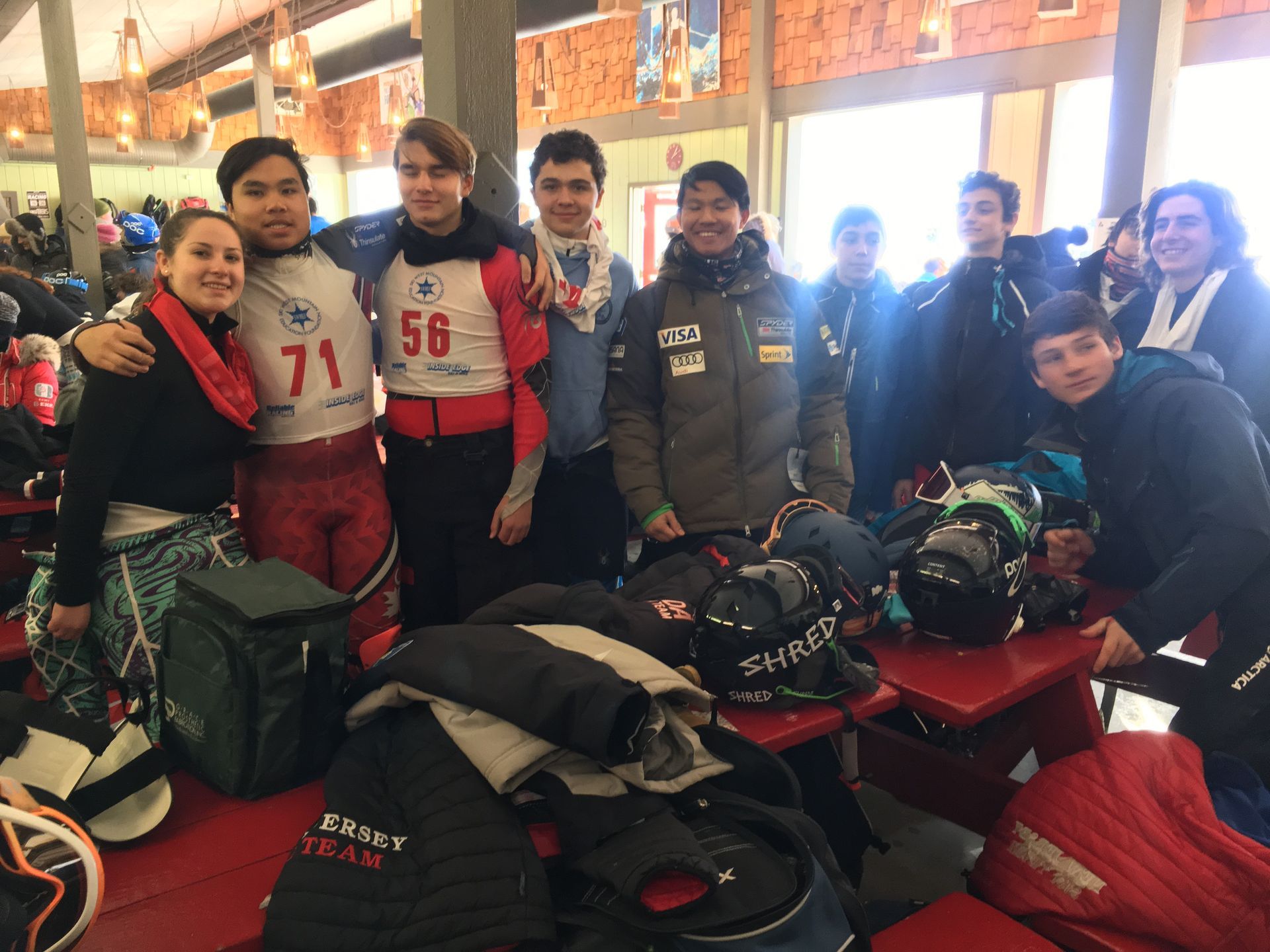 Group of people smiling, posing together. Inside a building, wearing winter clothing, with bags and gear in front of them.