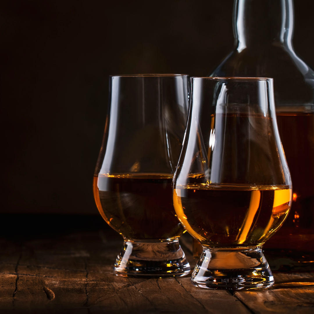 Two glasses of whiskey are sitting on a wooden table next to a bottle of whiskey.