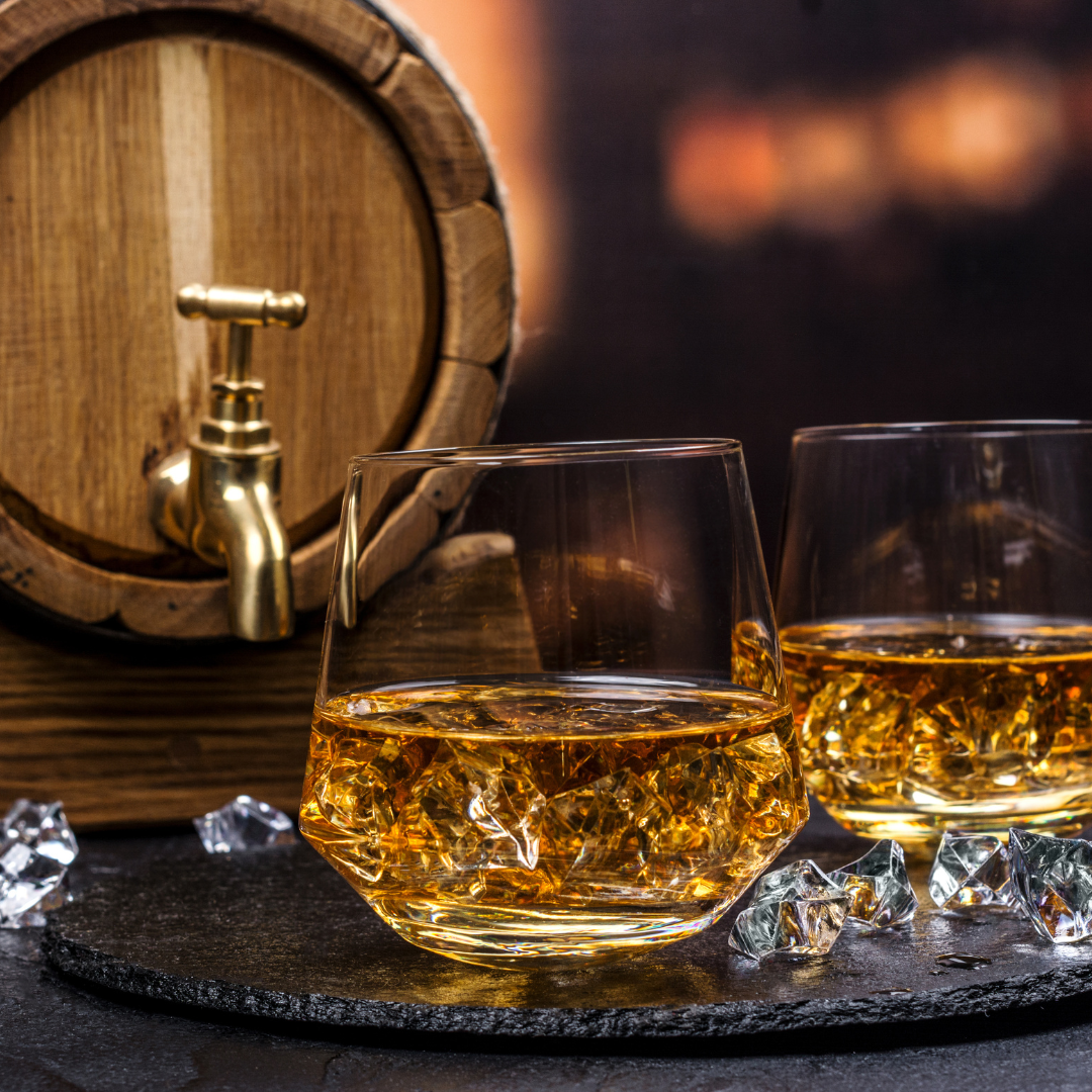 Two glasses of whiskey with ice in front of a wooden barrel.