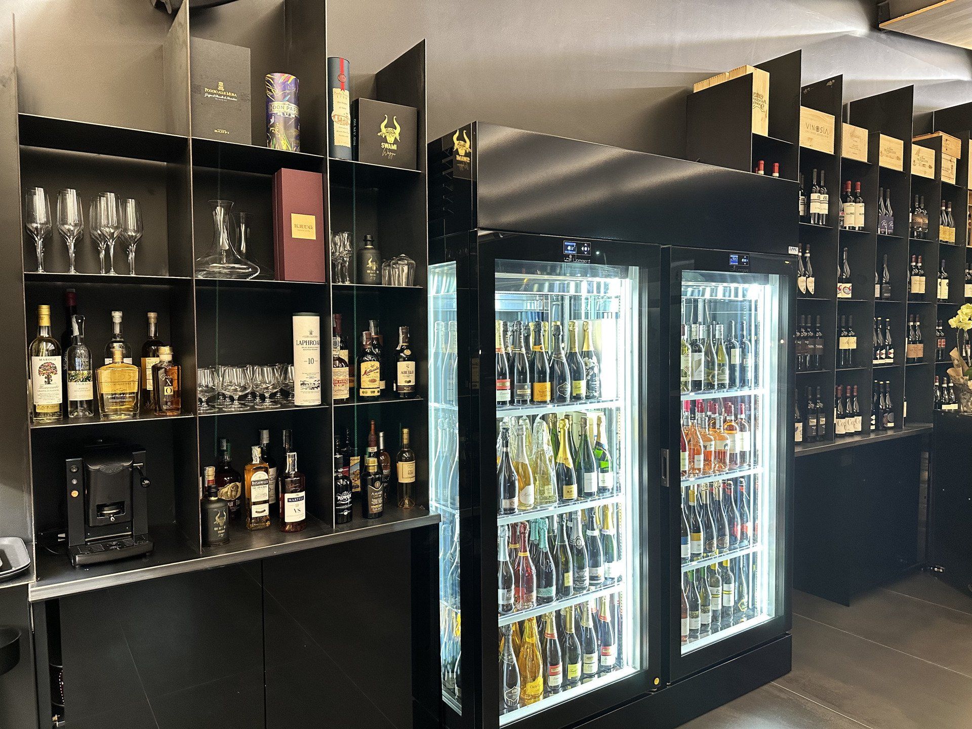 Scaffali e frigoriferi pieni di bottiglie di alcolici in un ambiente da bar.