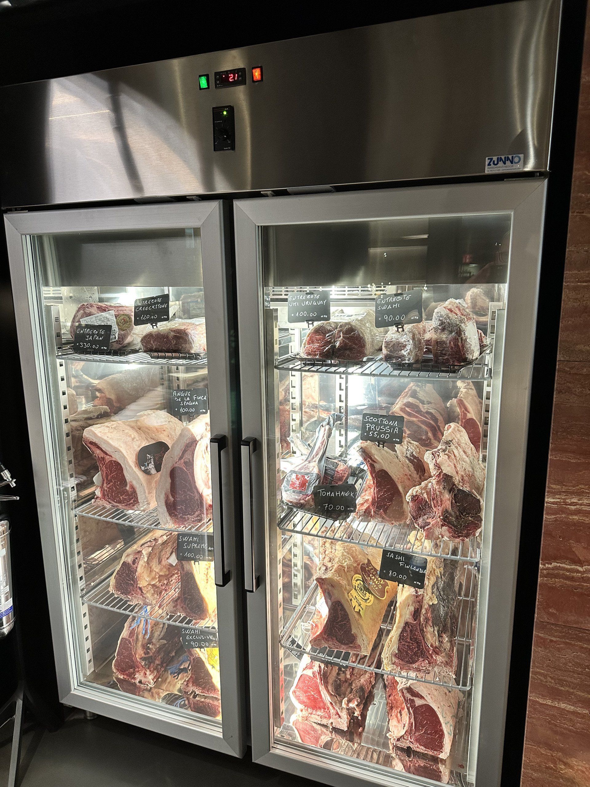 Un frigorifero commerciale espone vari tagli di carne rossa cruda dietro sportelli in vetro.