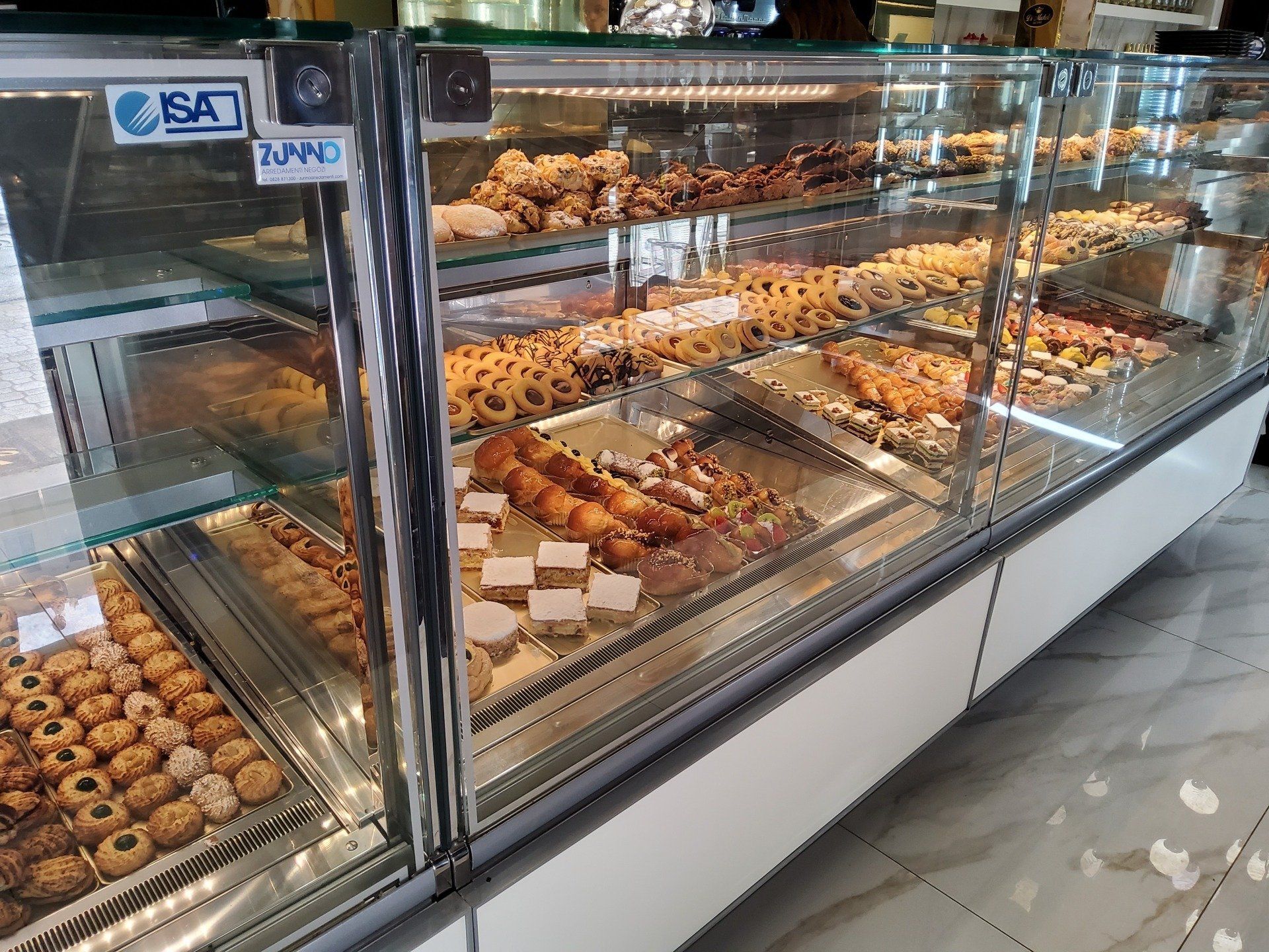 Vetrina da pasticceria con vari tipi di pasticcini e biscotti. Vetrina bianca e in acciaio inox con ripiani in vetro.