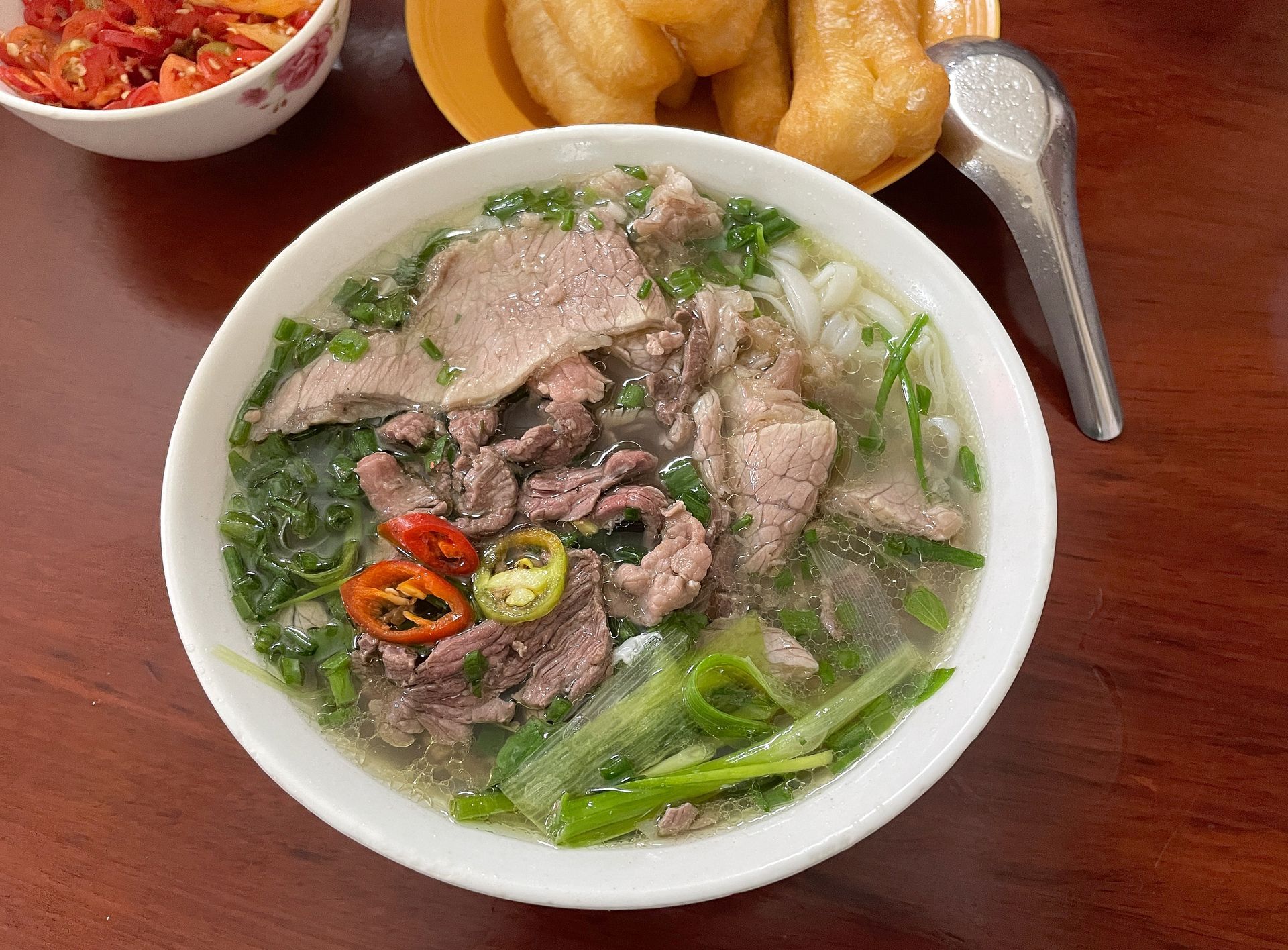 A steaming bowl of traditional Bat Dan beef pho is served with tender sliced beef, fresh scallions, and chili slices, accompanied by golden fried dough sticks.
