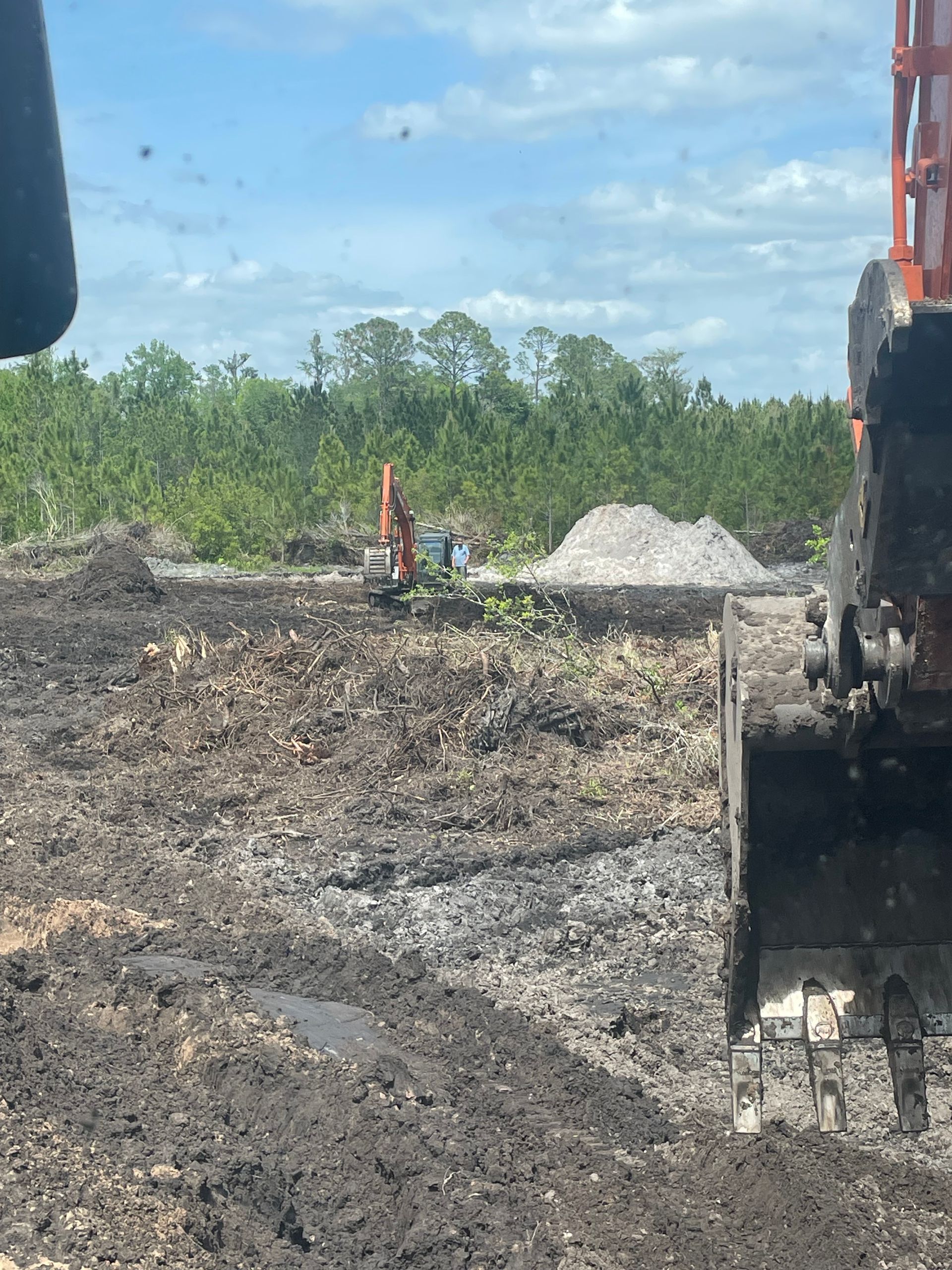 Excavator bucket at a muddy construction site with gravel piles and trees in the background