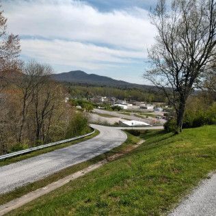 Winding road through a grassy hillside overlooking a small town and distant mountains under a cloudy sky