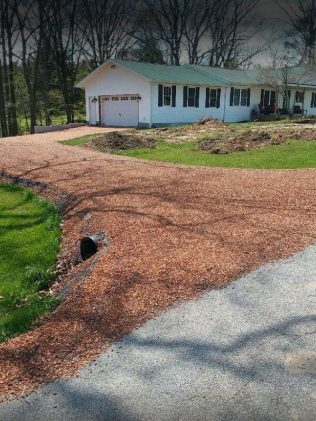 White house with green roof and attached garage beside a gravel driveway and grassy yard, with trees in back.