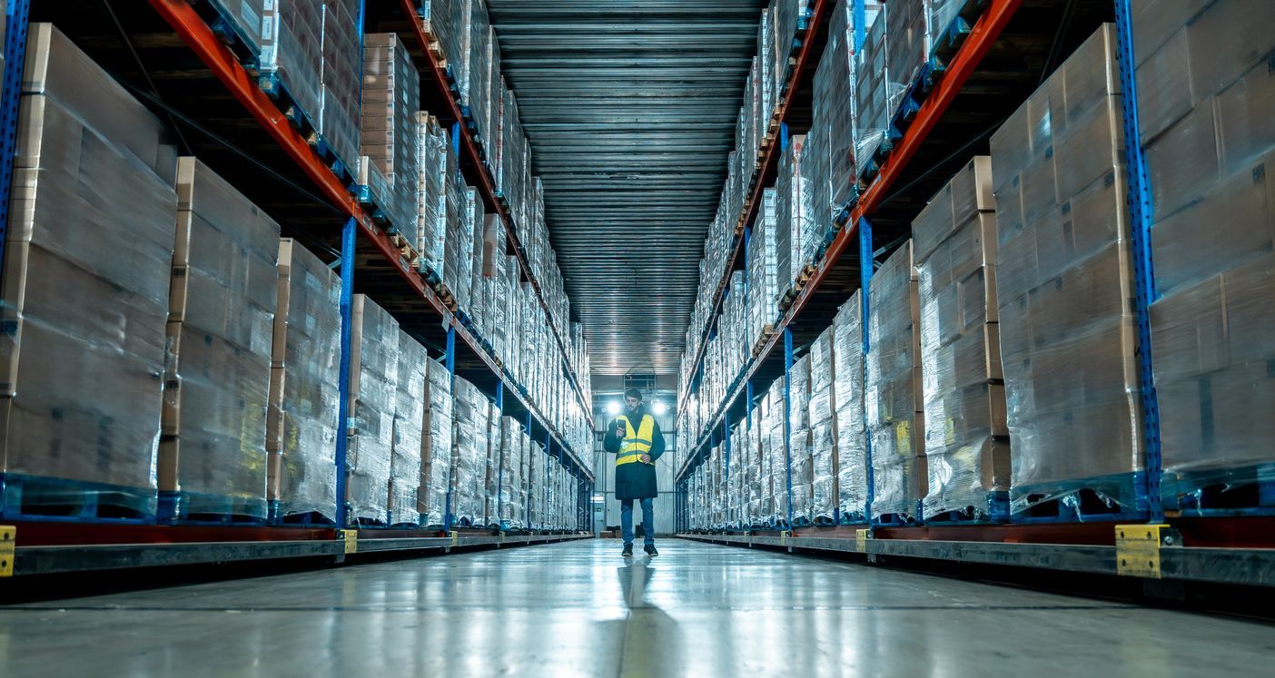 A warehouse aisle lined with stacked pallets and shelves, with a person in a reflective vest standing at the far end.