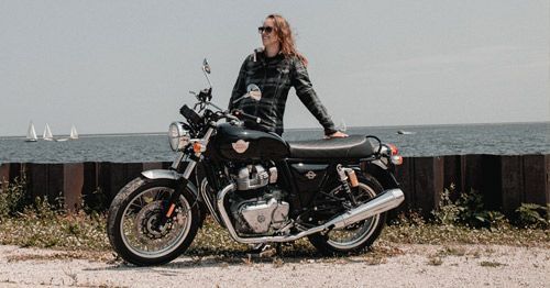 Pessoa vestida com roupa de couro preta em pé ao lado de uma motocicleta preta na costa.