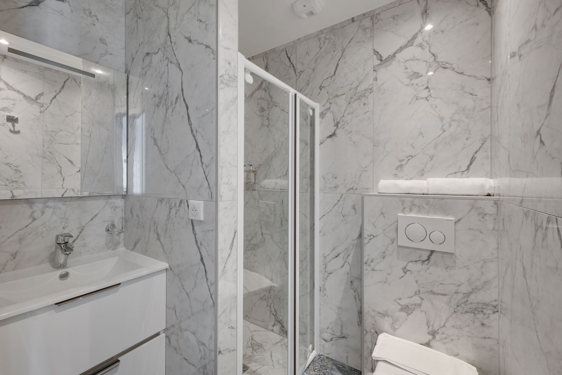 Marble-tiled bathroom with shower, sink, toilet. White walls, silver fixtures.