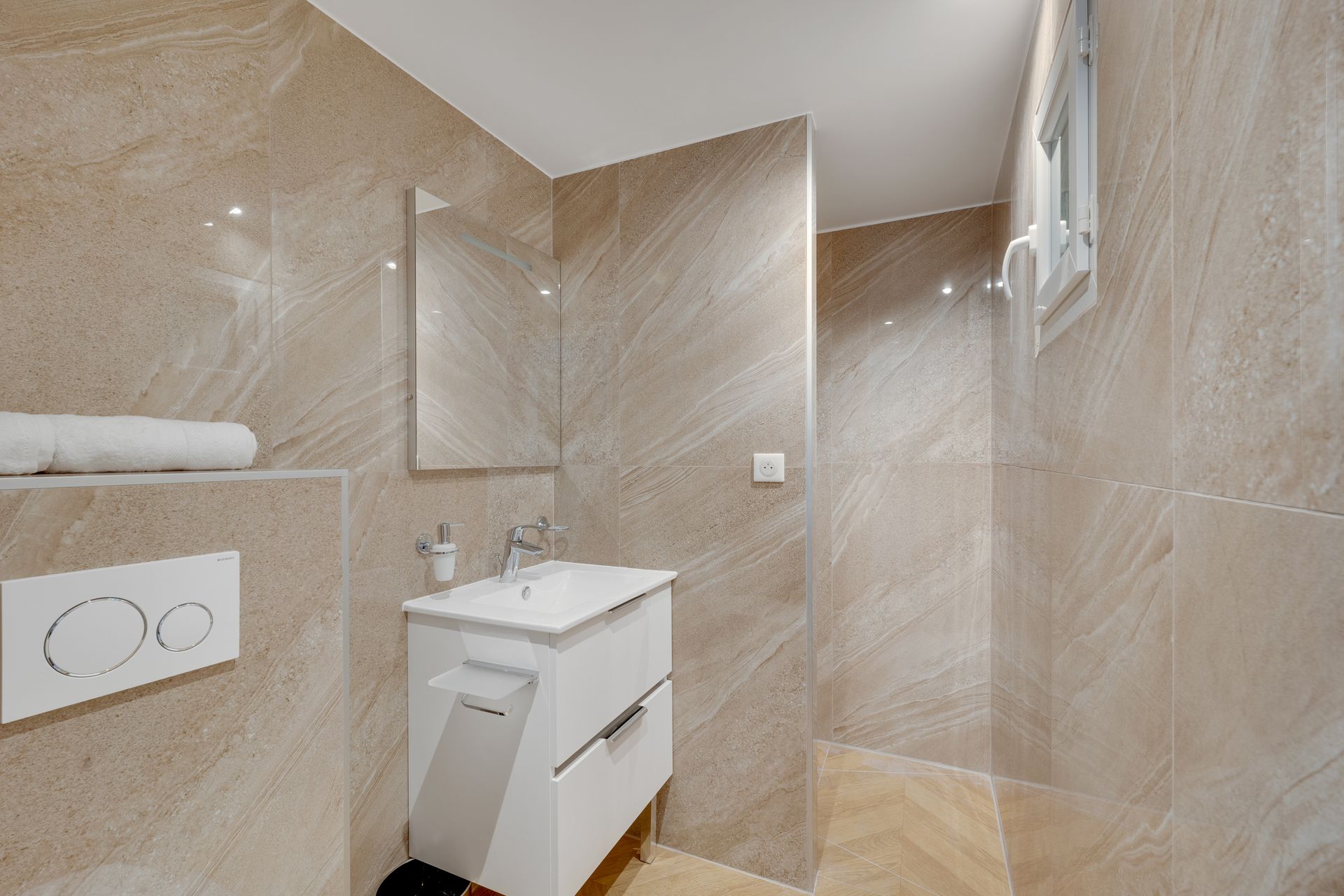 Small, beige-tiled bathroom with white vanity and window; a towel rests on a shelf.