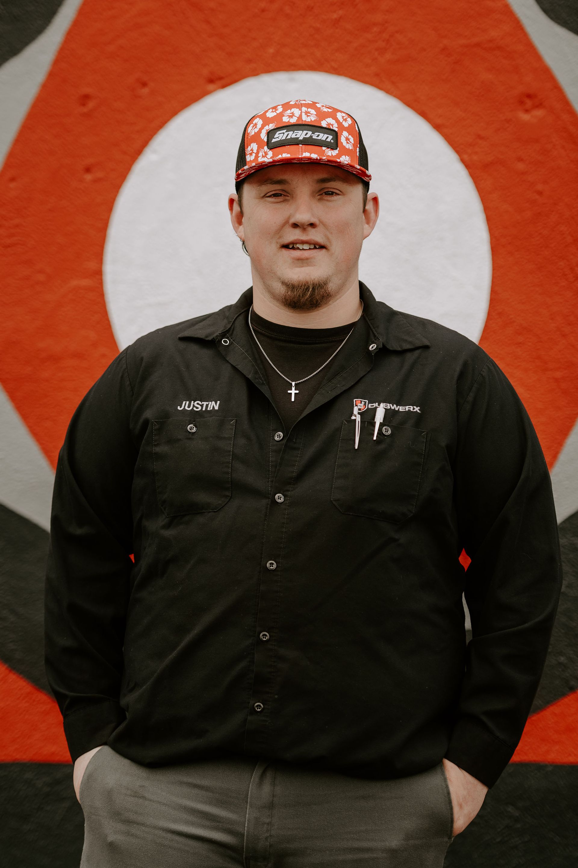 Person standing in front of an orange-and-white bullseye mural, wearing a black jacket and patterned cap.