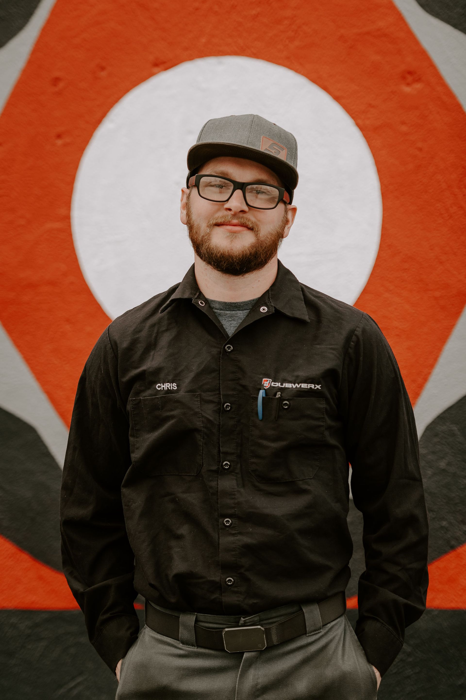 Man standing in front of a red-and-white circular sign, wearing glasses, a cap, and dark work clothes