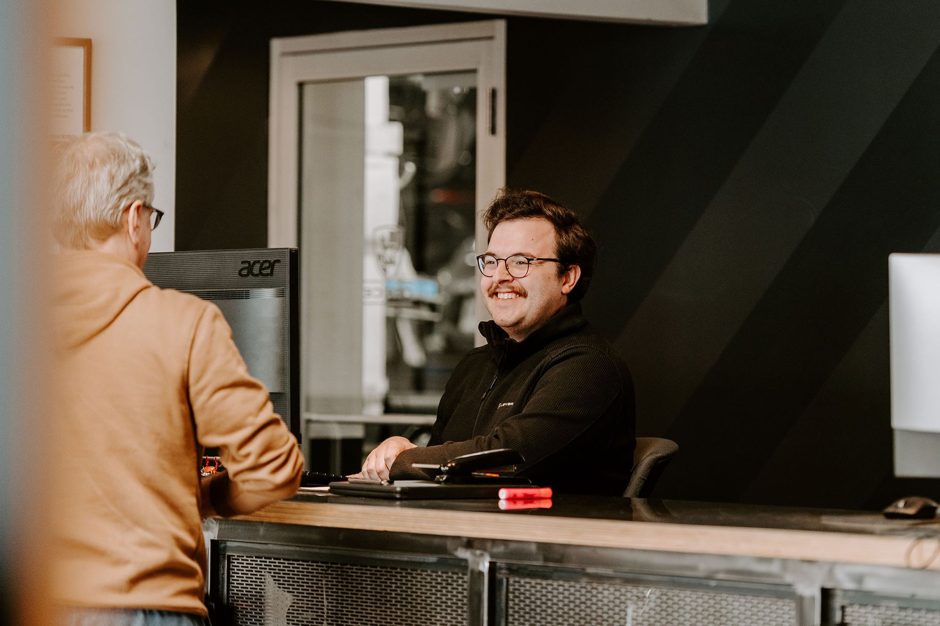 Two people talking across a front desk in a modern lobby, one seated with a laptop, one standing in a tan coat.