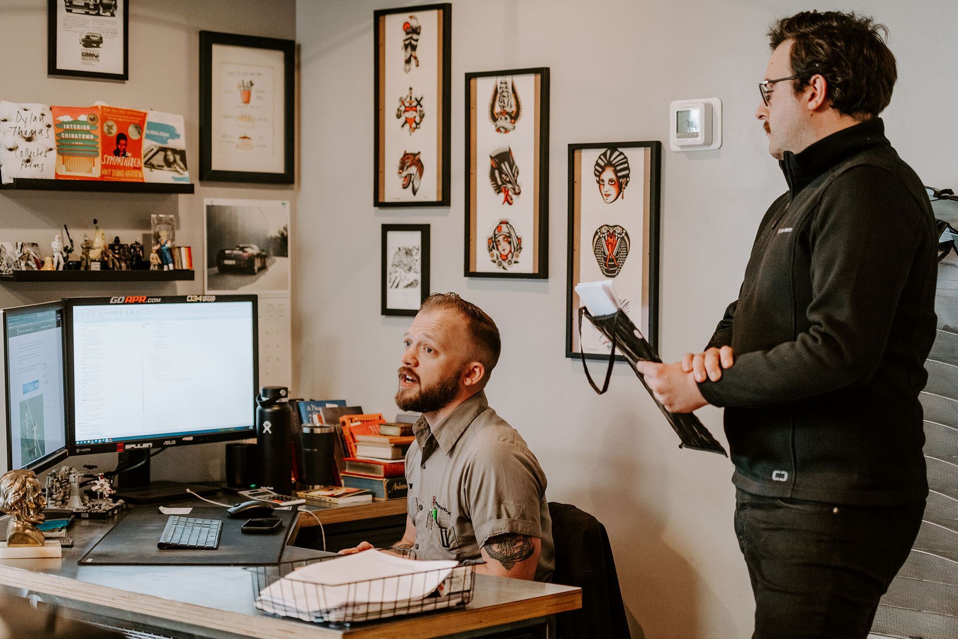 Two coworkers in an office, one seated at a computer and one standing with a tablet.