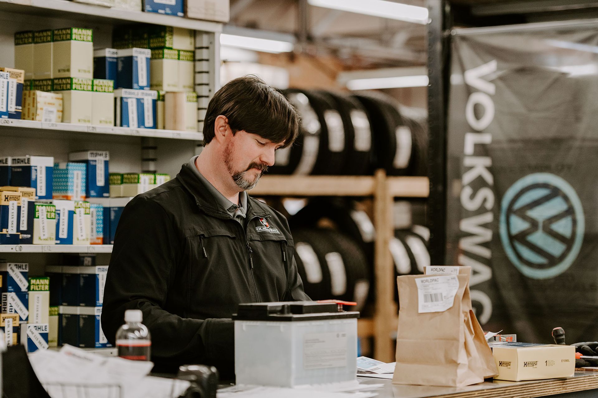 Man in a workshop, working at a counter beside shelves of supplies and a Volkswagen banner.