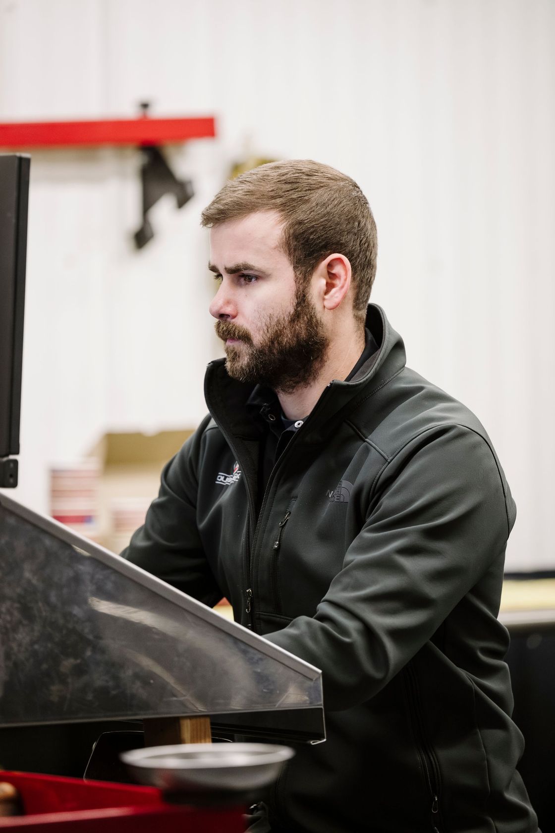 Man in a black jacket working at a desk or workstation, seen in side profile indoors