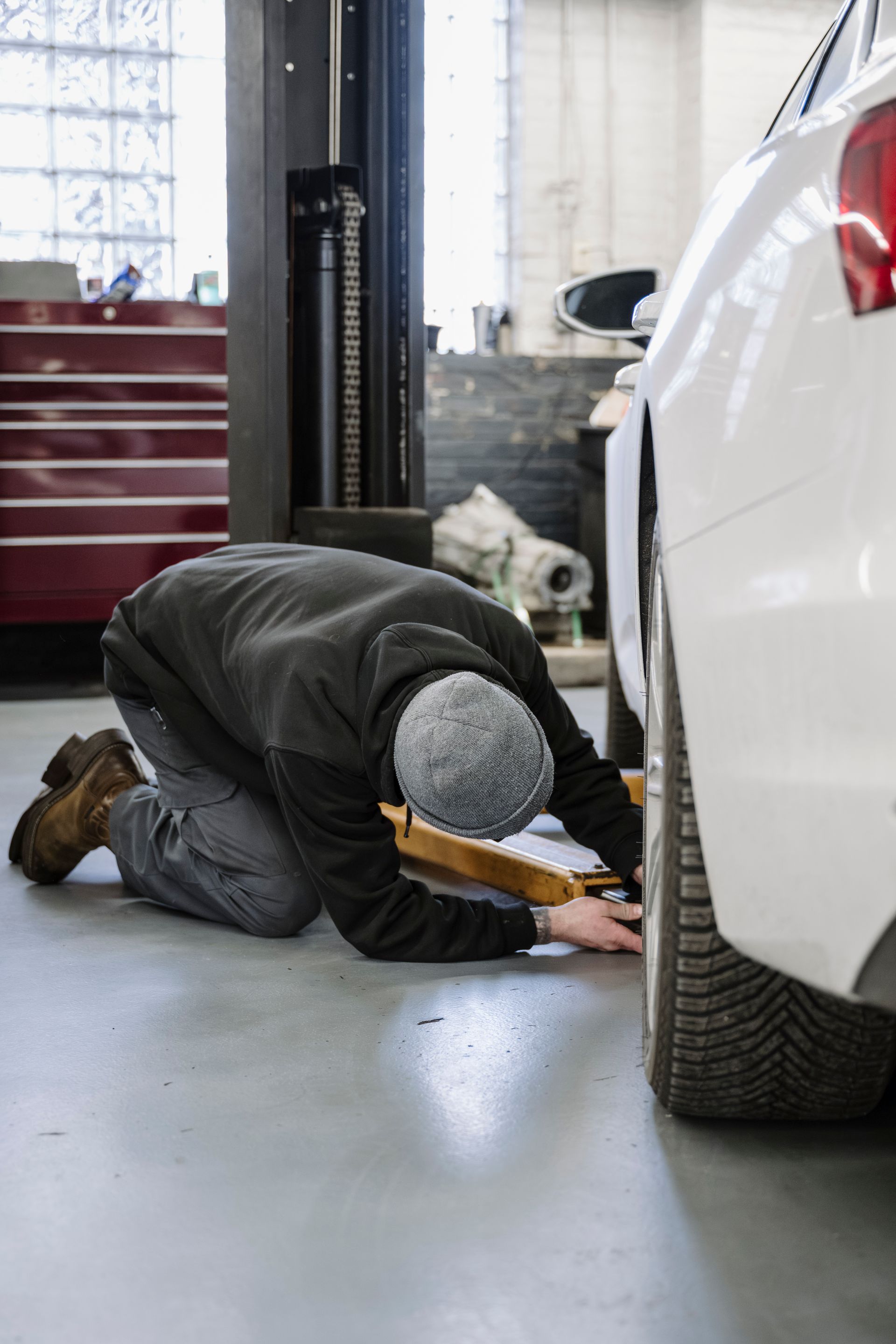 Mechanic kneeling beside a white car, inspecting the front wheel in a garage.