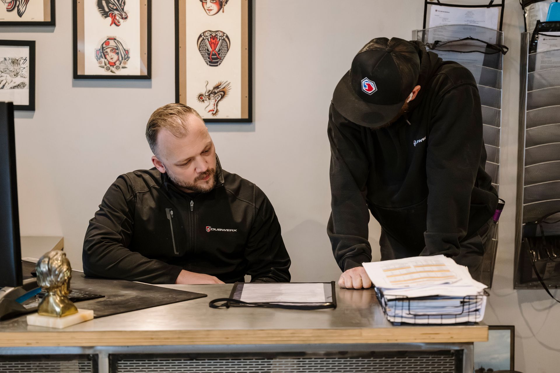 Two people at a desk reviewing paperwork in an office, with framed artwork on the wall behind them.