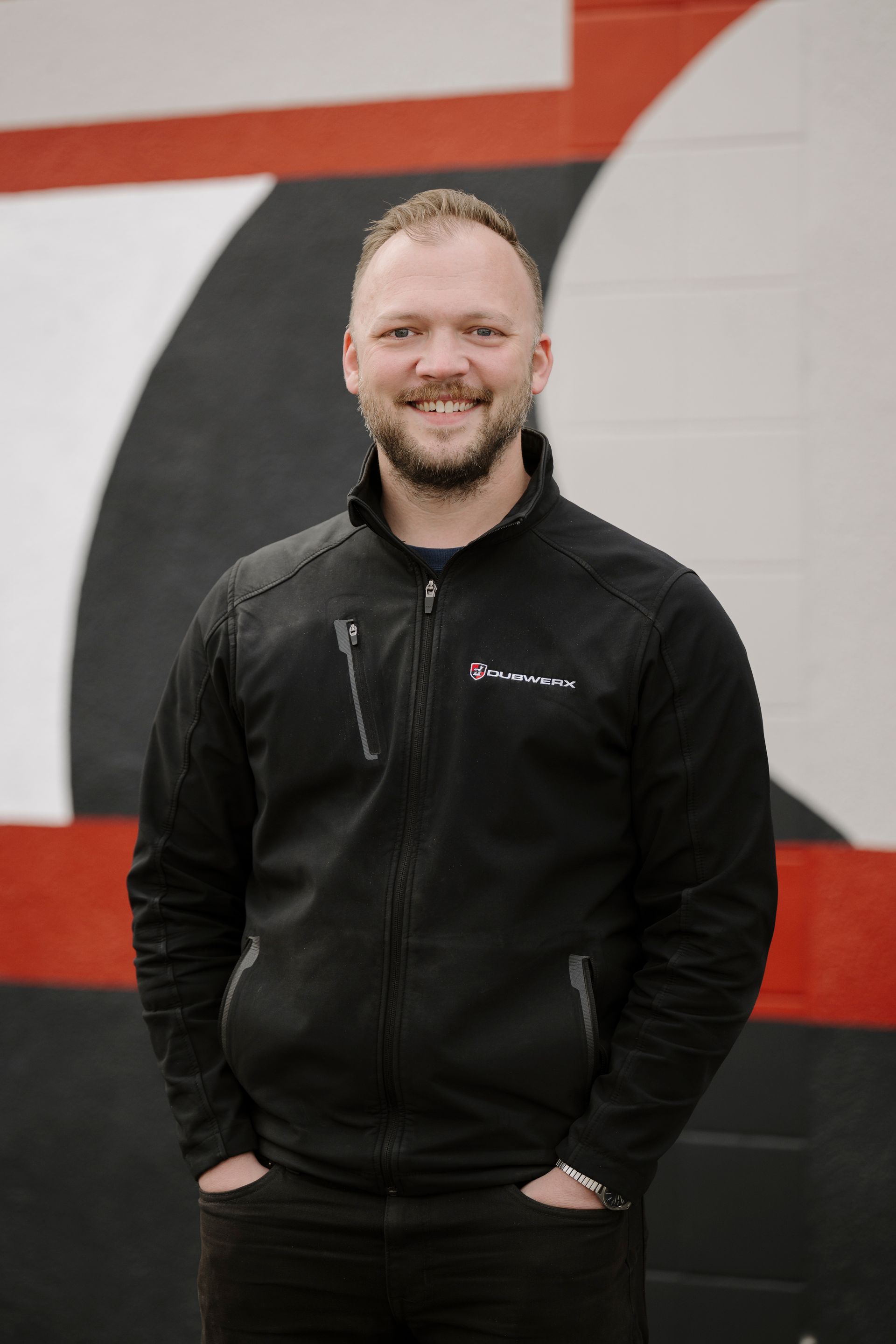 Smiling man in a black jacket standing in front of a red, black, and white wall