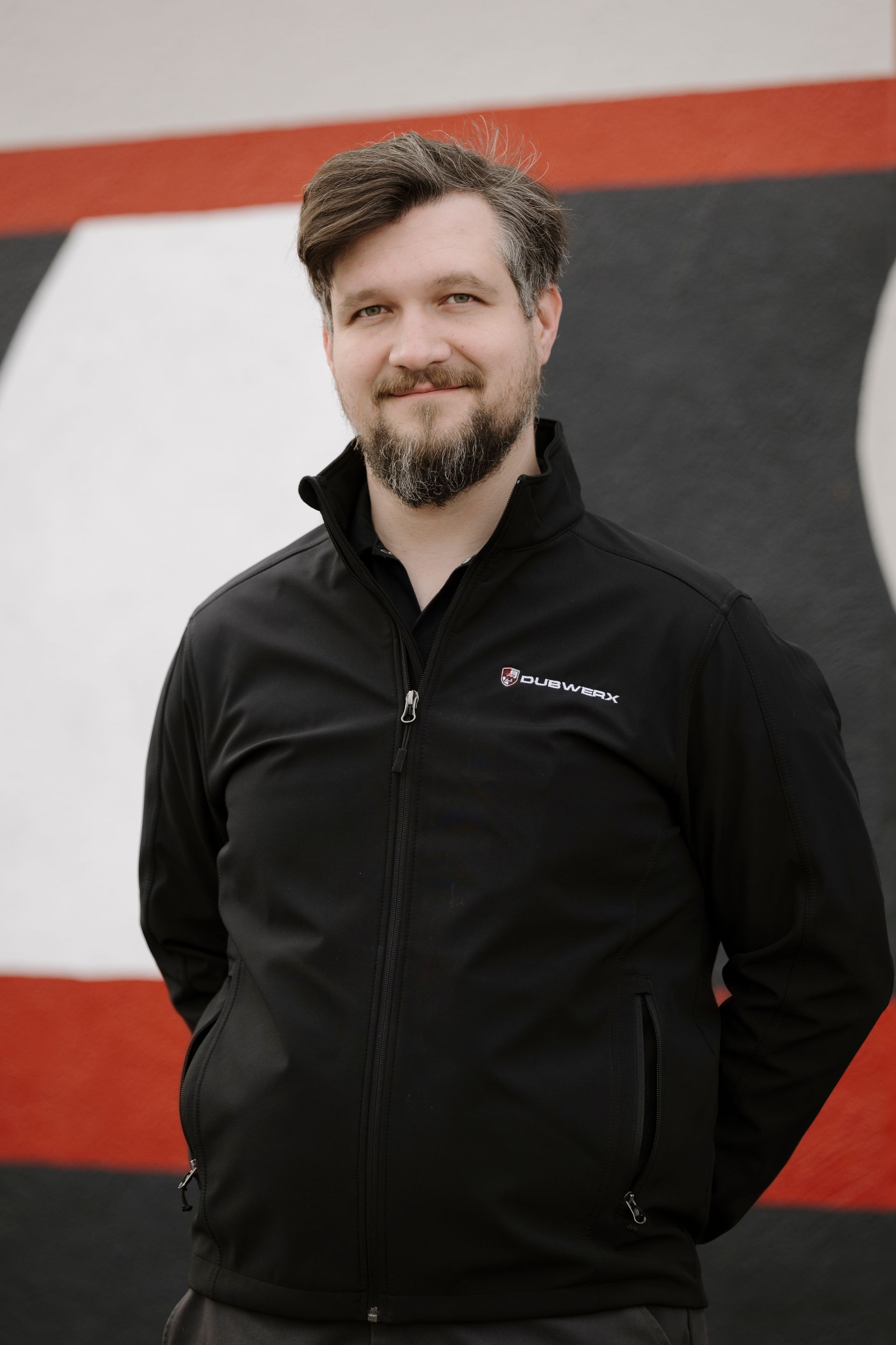 Man in a black jacket standing with hands behind his back against a red, black, and white mural