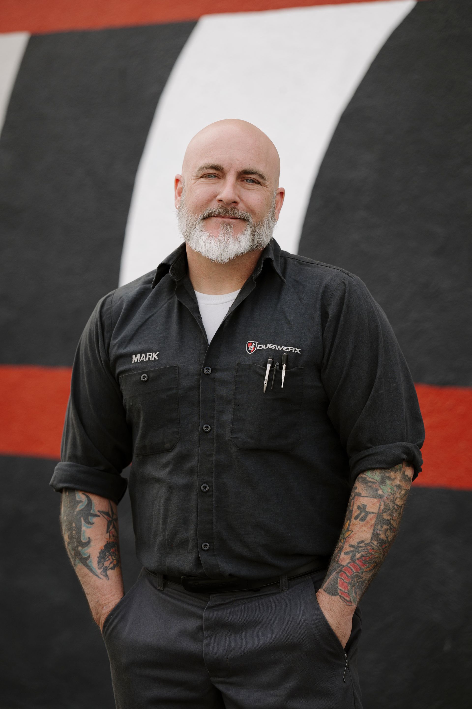 Bearded man in a black shirt with tattooed arms, standing against a red, white, and black mural.