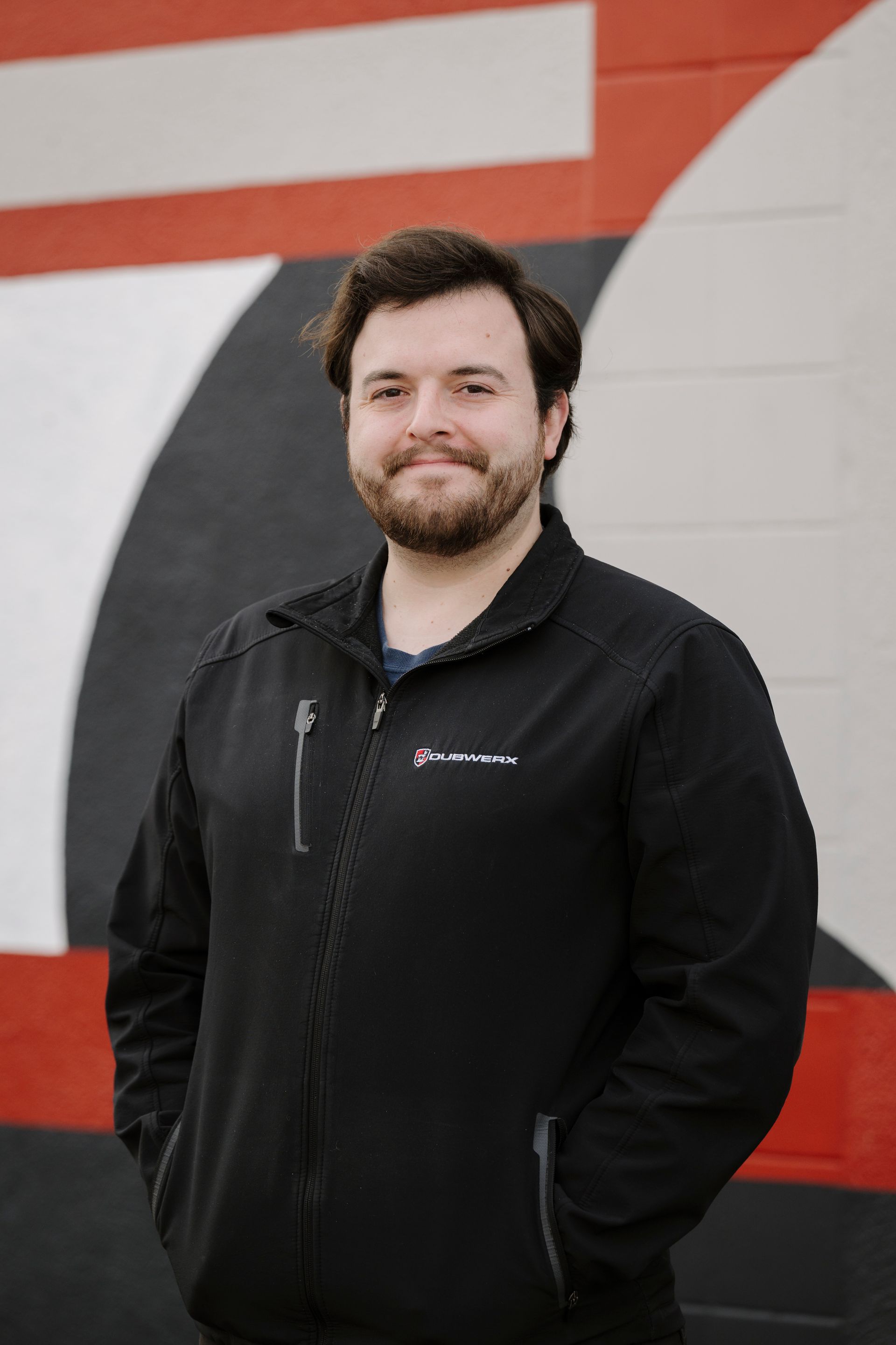 Man in a black jacket standing against a red, white, and black mural, smiling at the camera