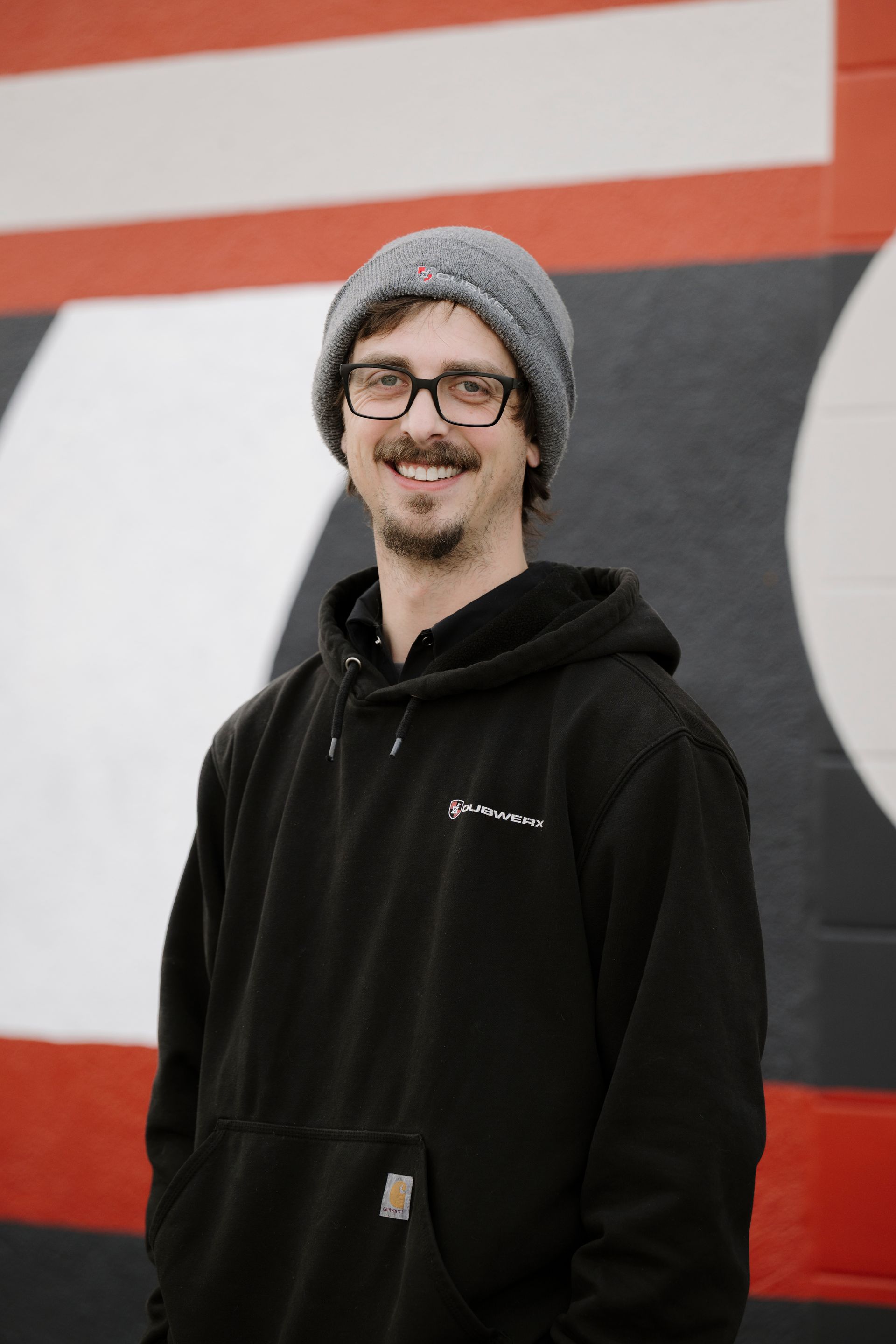 Person in a gray beanie and black hoodie smiling in front of a red, white, and black wall.