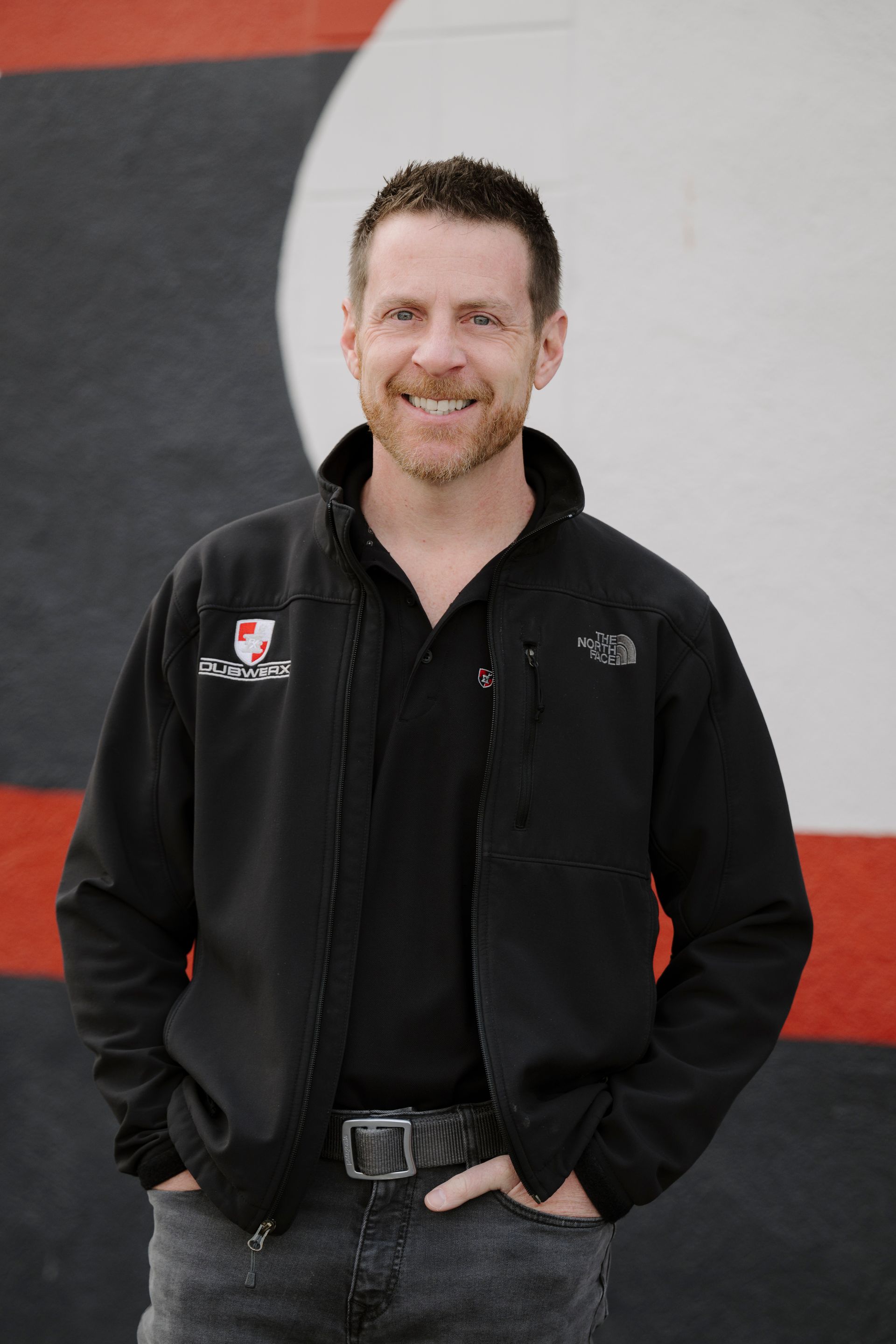 Smiling man in a black jacket standing with hands in pockets against a gray and red wall