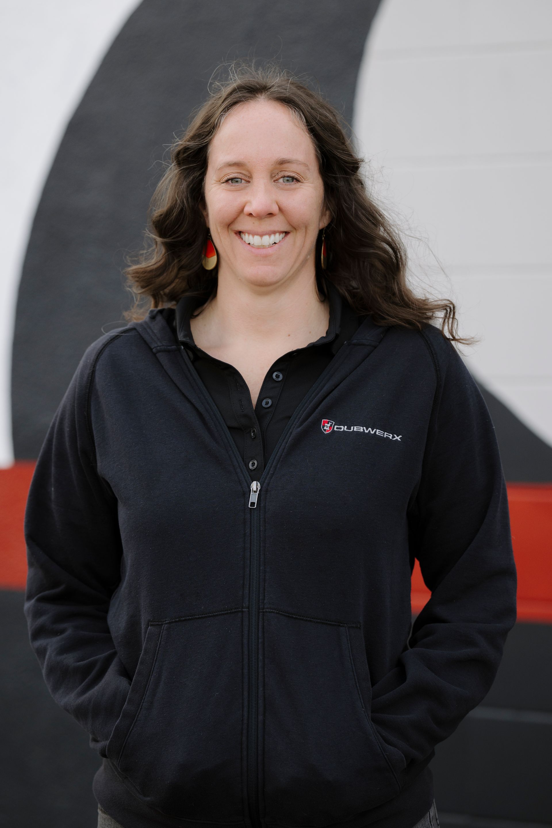 Person smiling in a black jacket standing outdoors in front of a black, white, and red wall