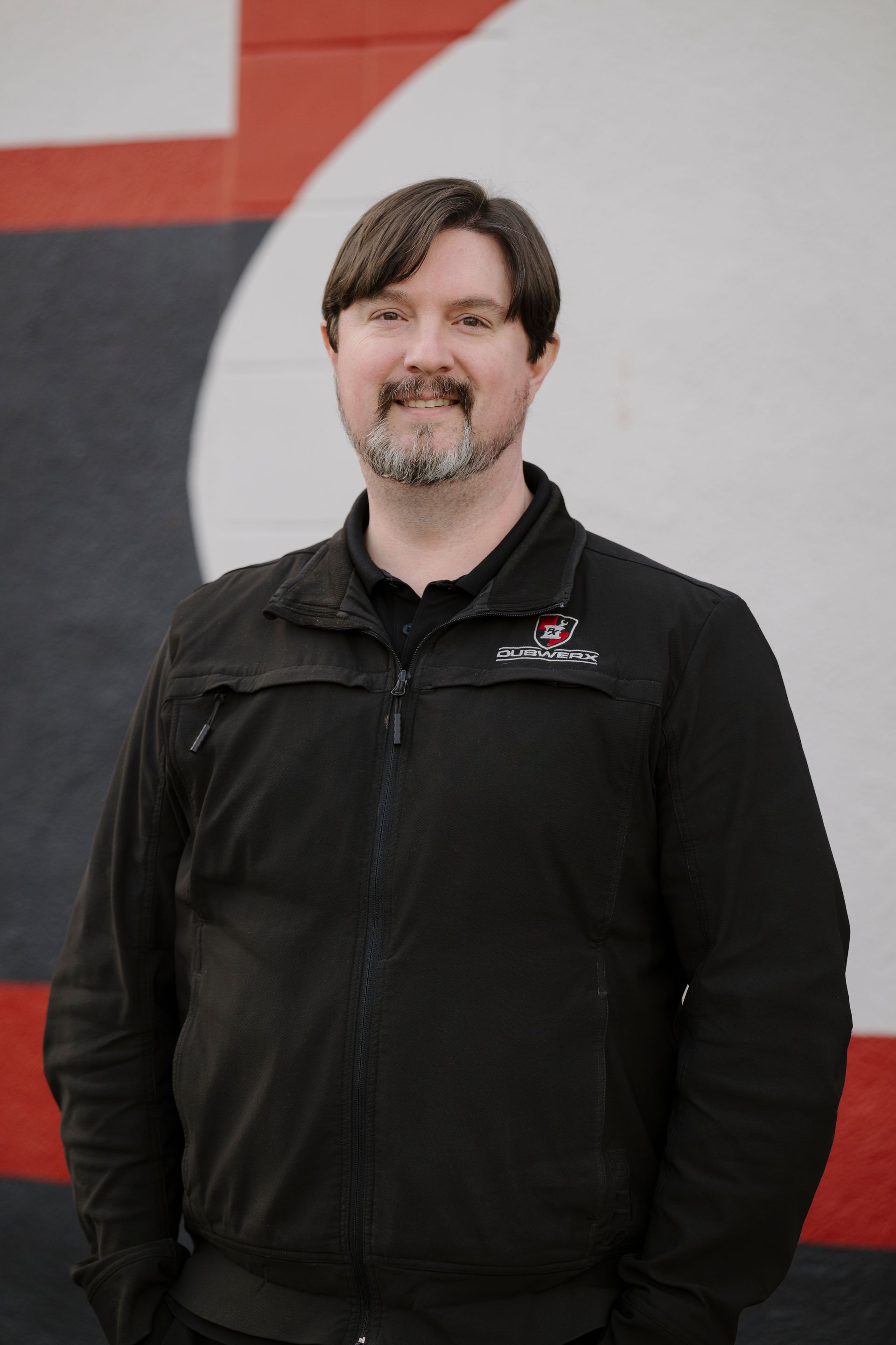 Person in a black jacket standing against a white and red wall, smiling softly.