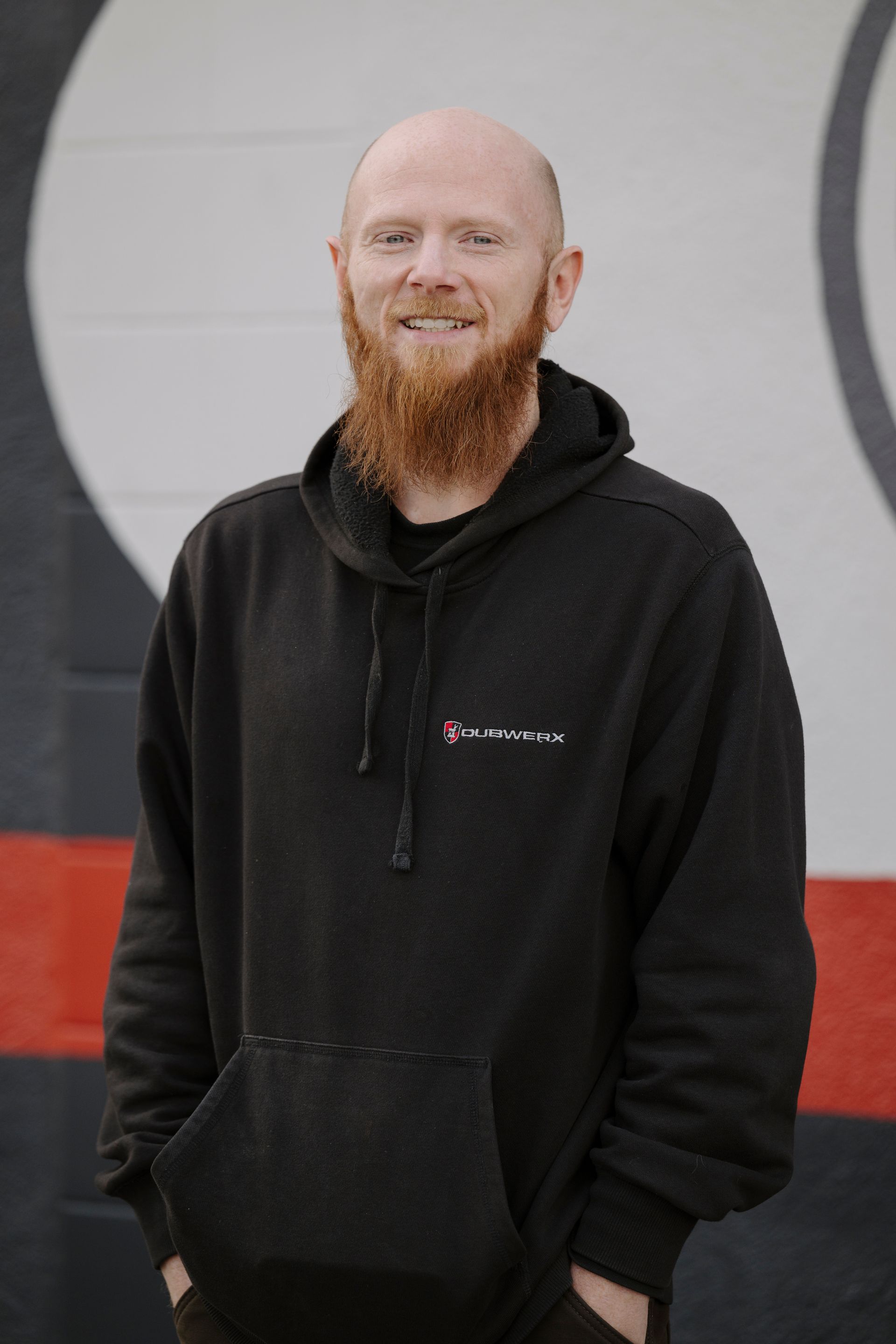 Smiling bald man with a red beard wearing a black hoodie, standing against a gray and red wall.