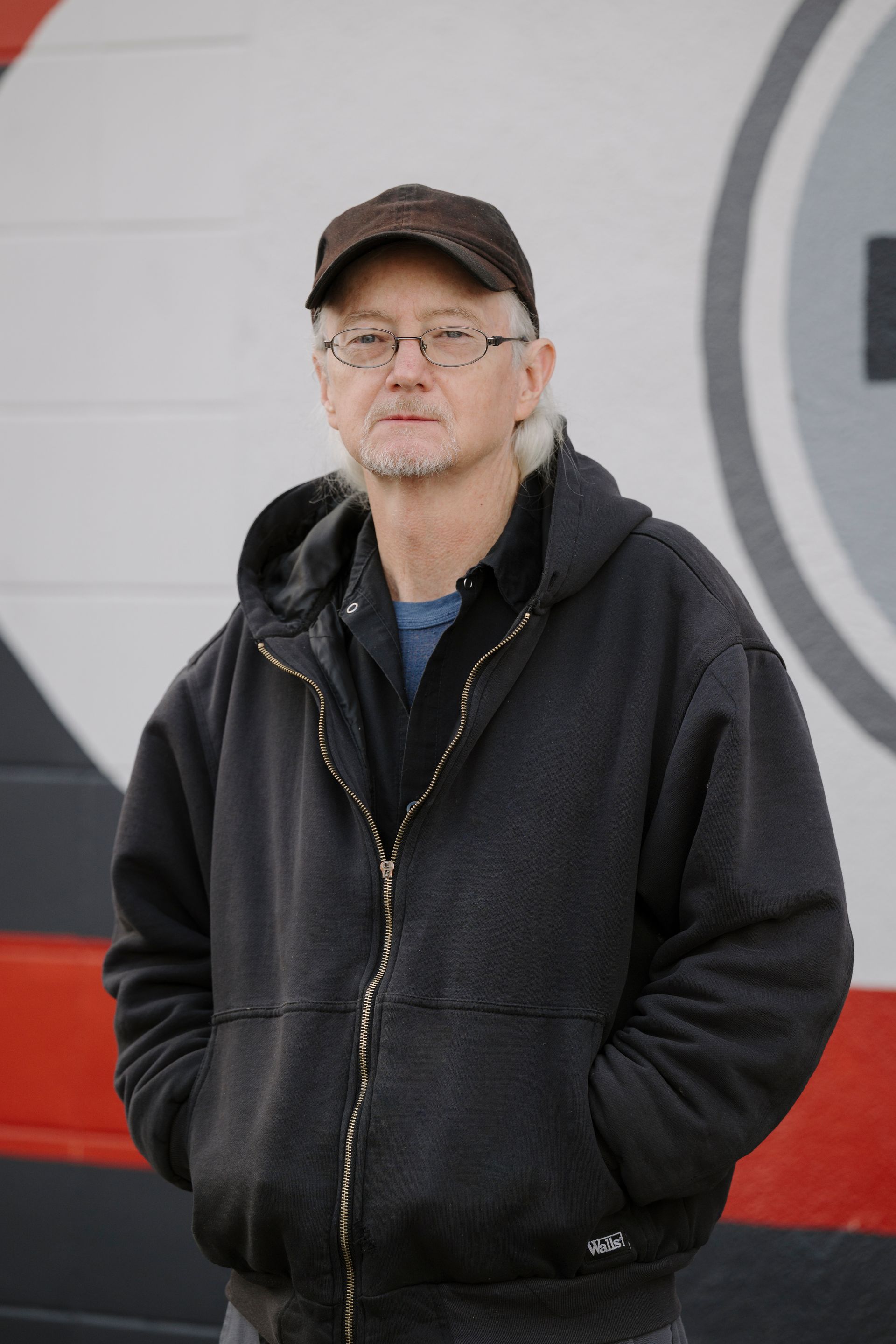 Person in a black hoodie and cap standing outdoors against a red-and-white wall.