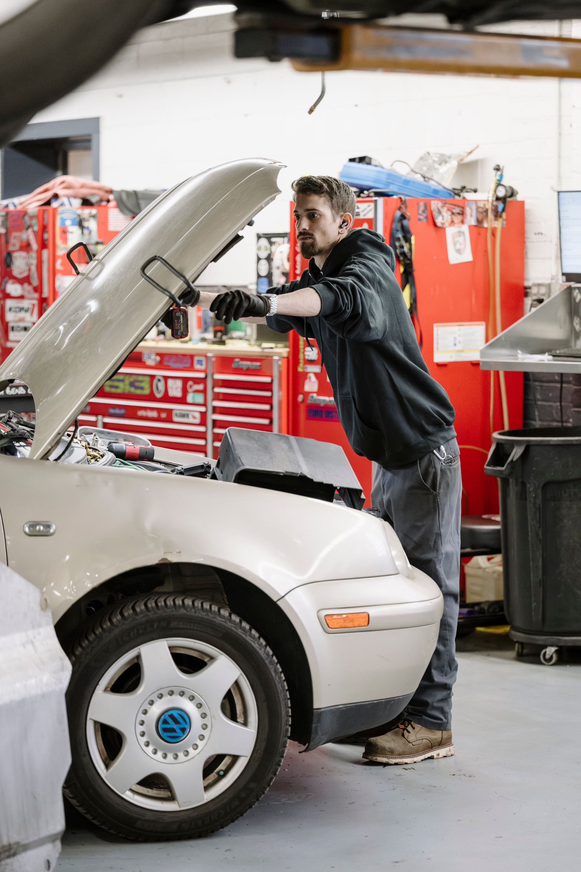 Mechanic working under the hood of a white car in an auto shop with red tool cabinets.