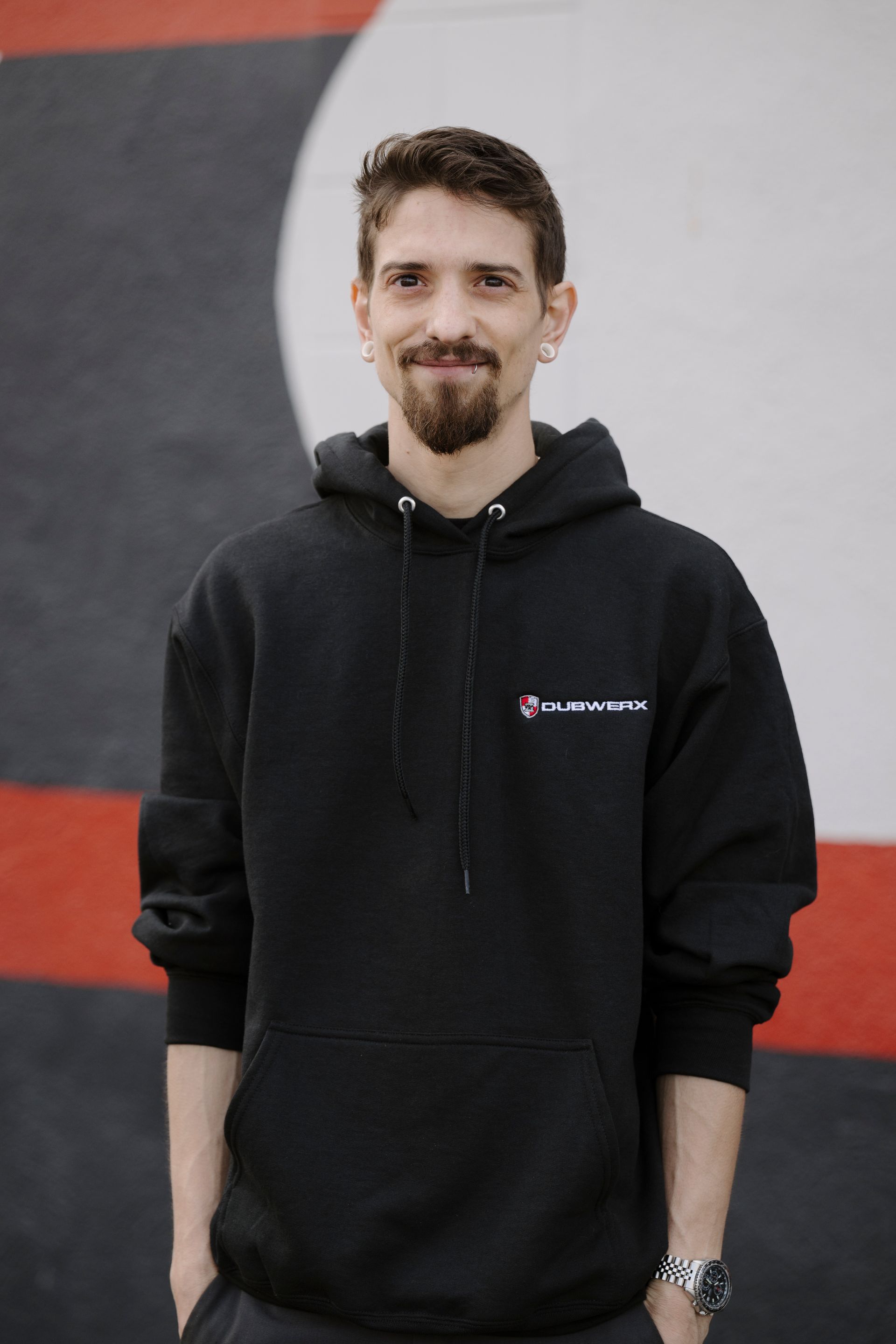Person in a black hoodie standing against a red, gray, and white wall, smiling at the camera.