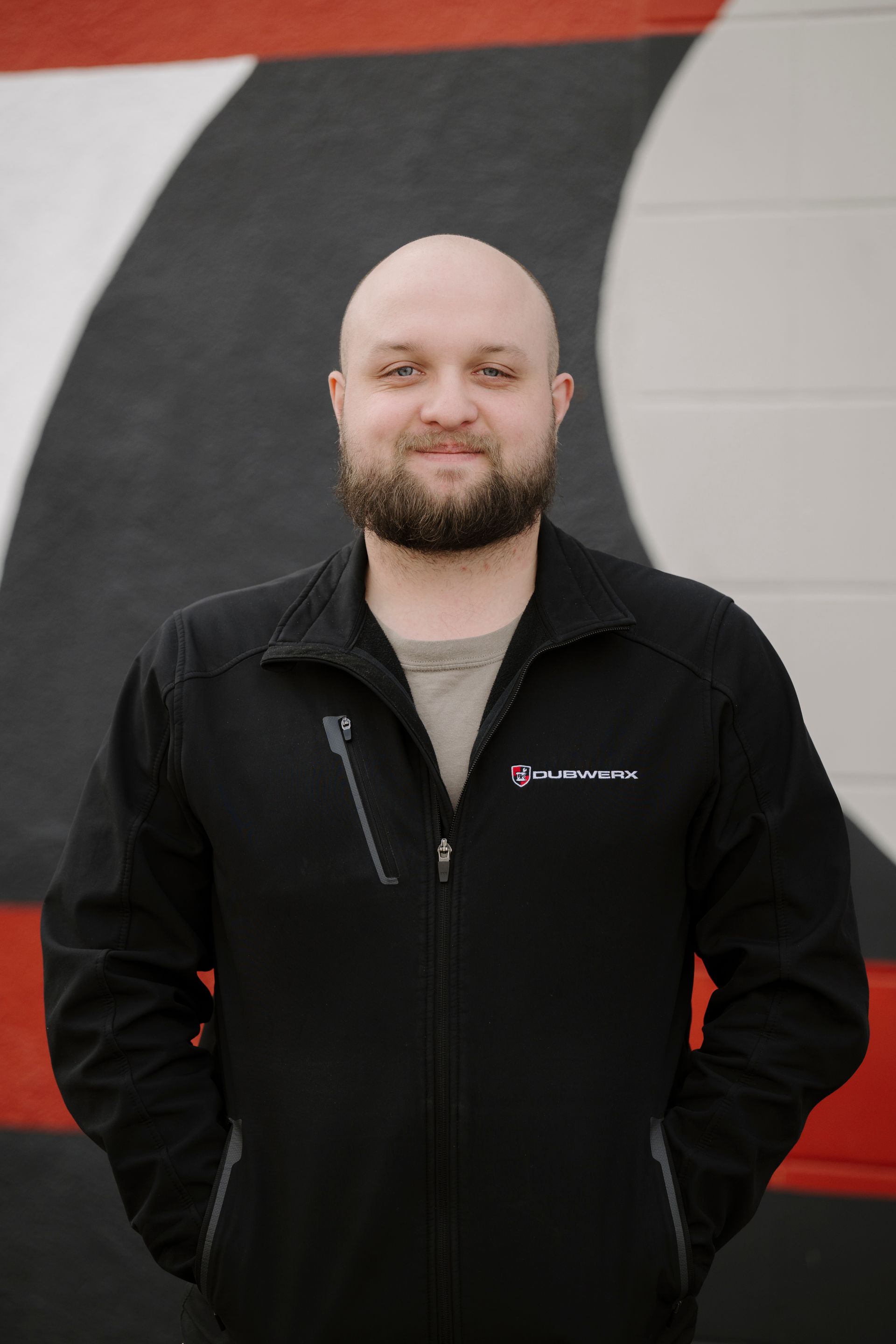 Smiling bald man in a black jacket standing in front of a black, red, and gray wall