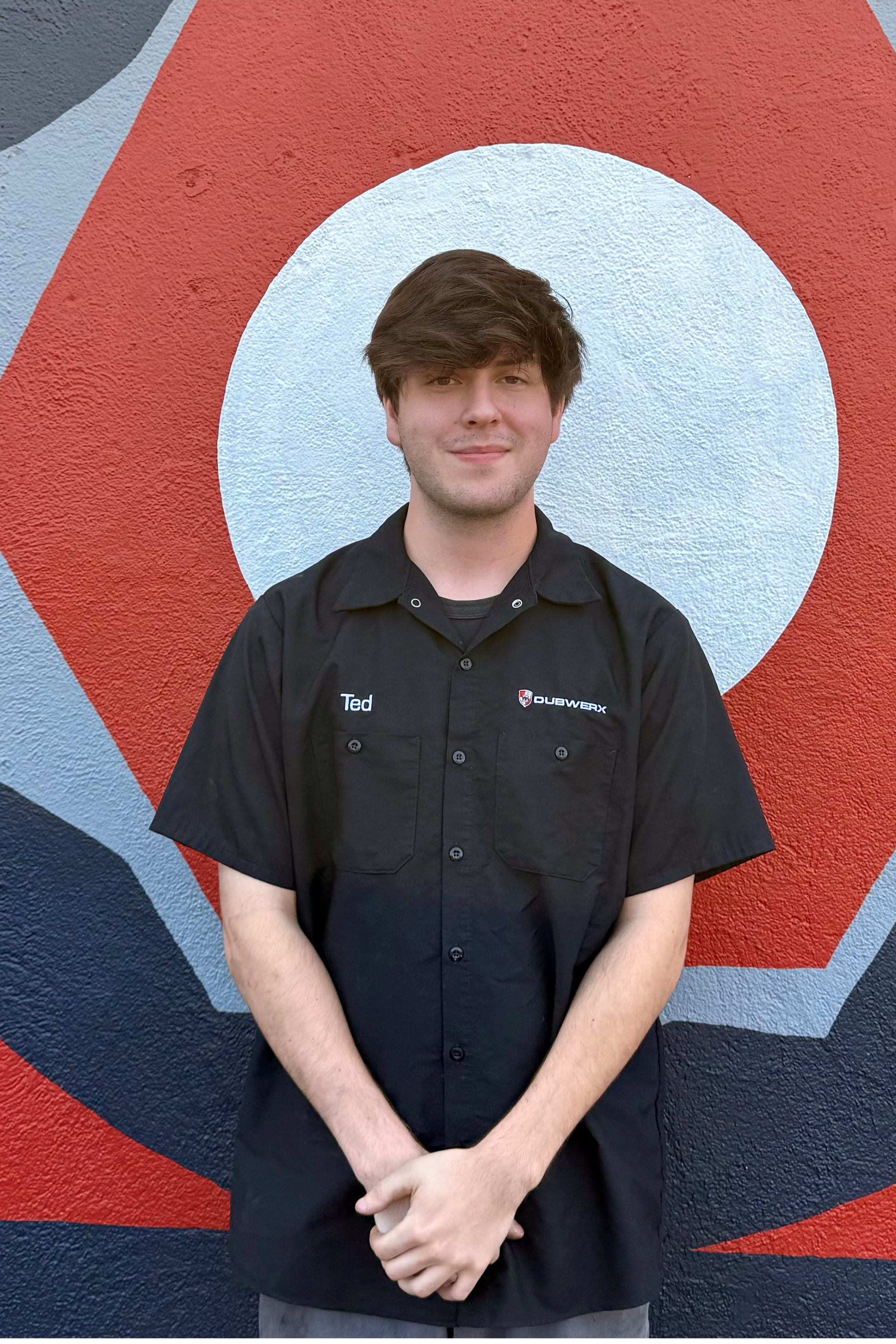Smiling man in a black polo shirt standing against a wooden plank wall.