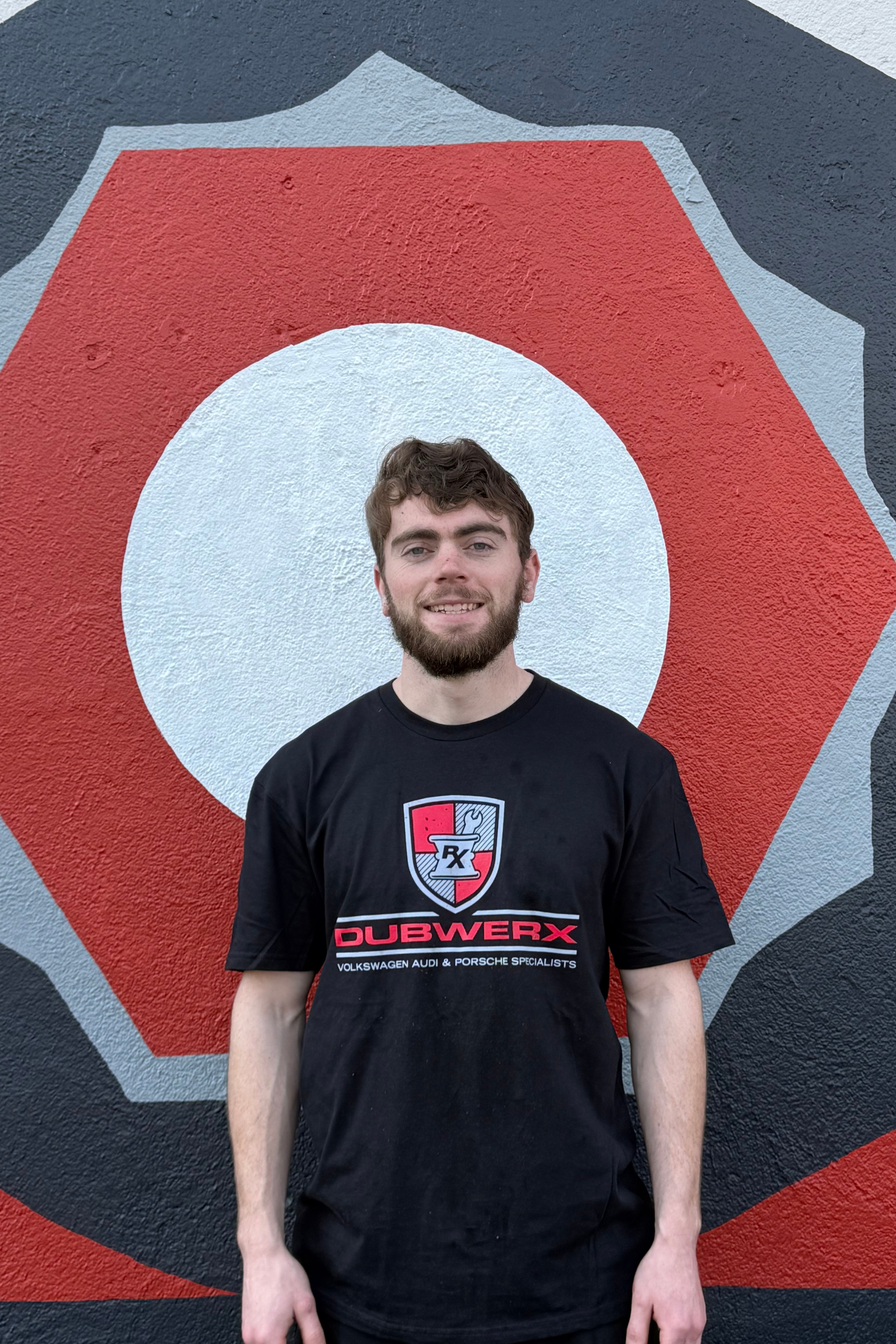 Man standing in front of a red hexagon mural with a white circle, wearing a black T-shirt and smiling.