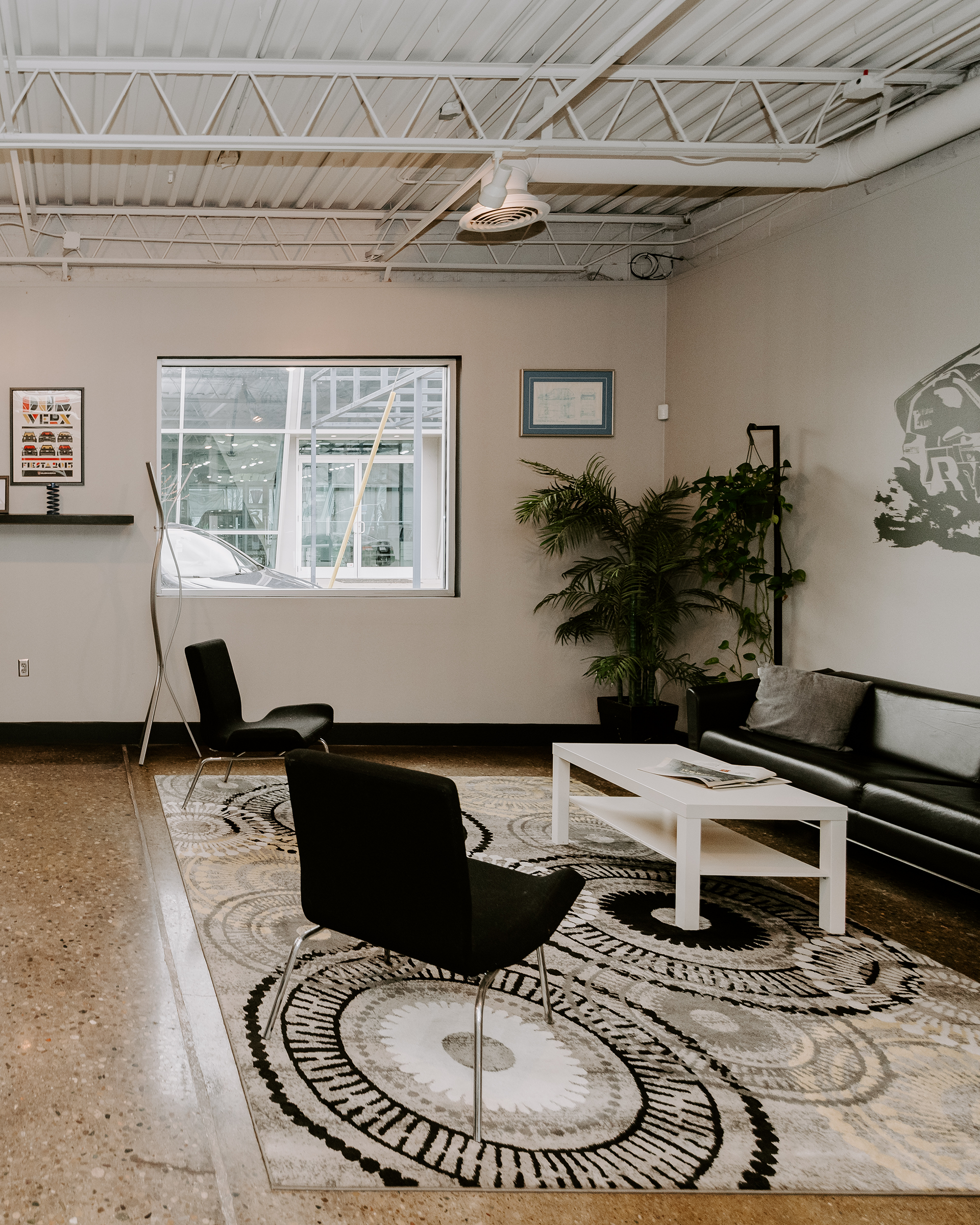 Modern lounge with black chairs, white coffee table, patterned rug, plants, and large window.
