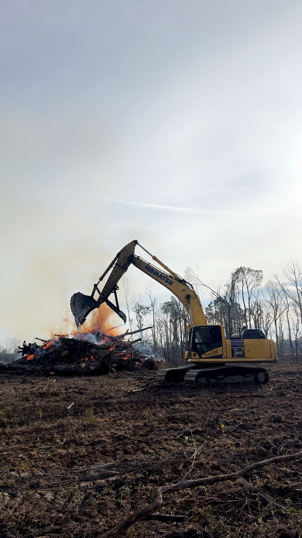 Excavator demolishing a burning pile of debris in a field under a gray sky.