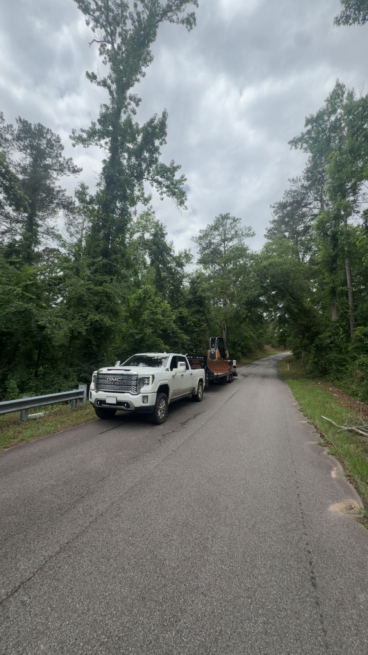 White pickup and orange utility vehicle parked on a narrow gravel road beside dense trees under cloudy skies