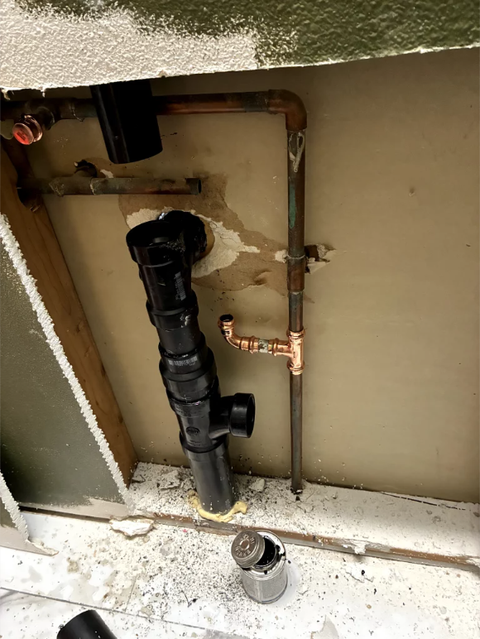 Exposed black and copper plumbing pipes inside a wall cavity with construction debris on the floor