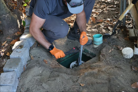 Worker in orange gloves inspecting a square pit with a metal rod at an outdoor excavation site