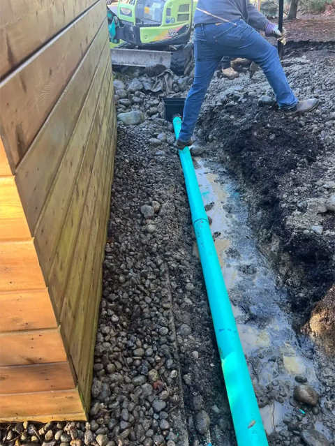 Worker beside an exposed turquoise pipe and excavator near a wooden building