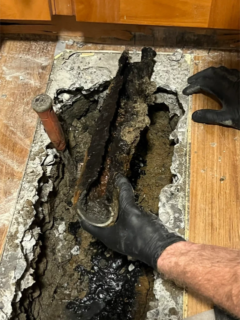 Gloved hands lifting a corroded pipe from a broken floor opening with black sludge debris