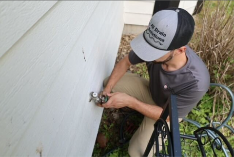 Man adjusting an outdoor water spigot beside a white house wall and metal railing
