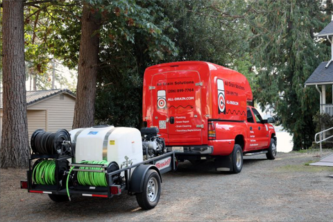 Red utility van towing a small trailer with tanks and hose reels on a gravel driveway in a wooded area